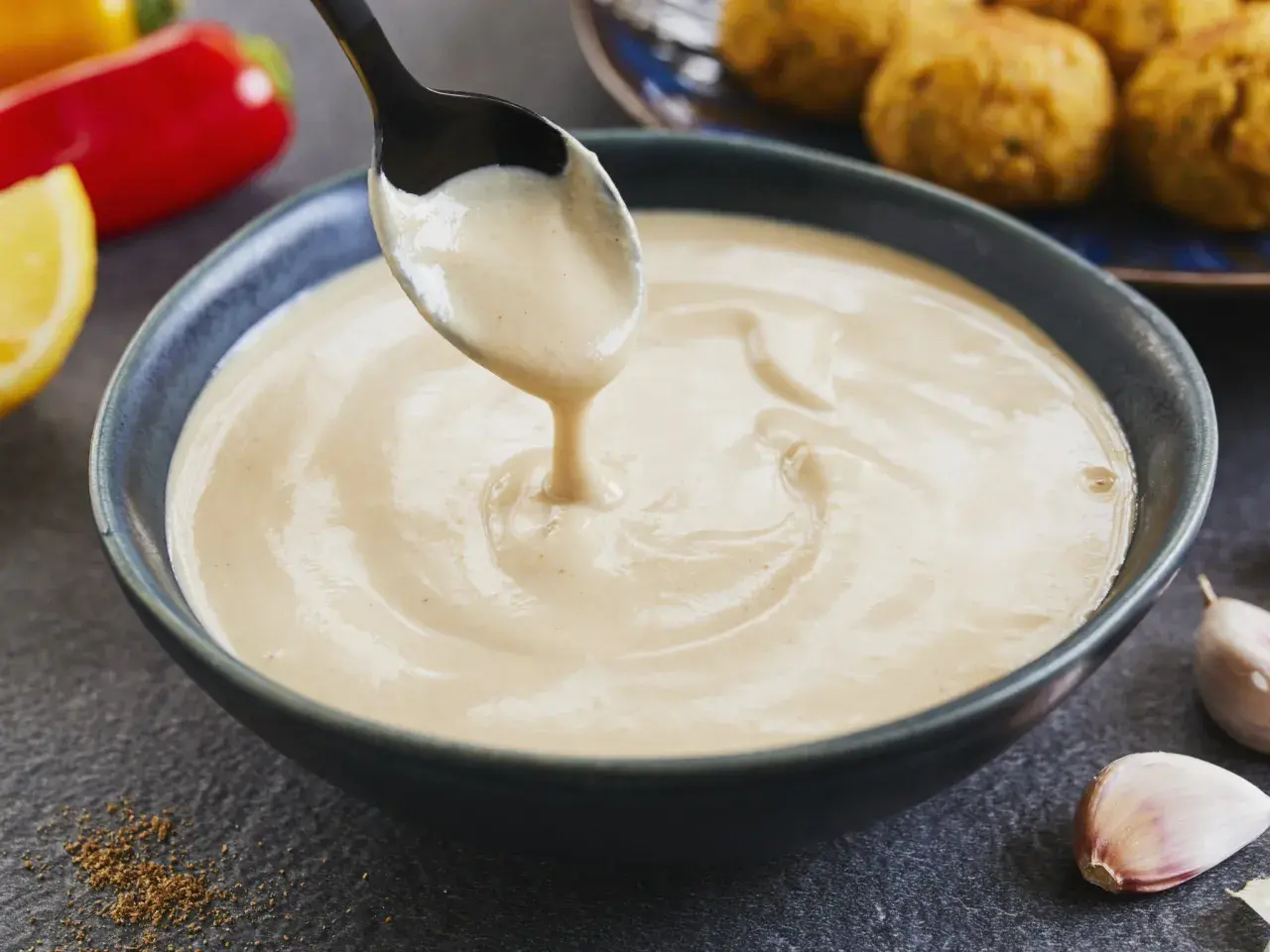 Cremige Tahini-Sauce tropft von einem Löffel in eine Schüssel. Sie ist die perfekte Begleitung zu Falafel, wie sie im Hintergrund zu sehen sind.