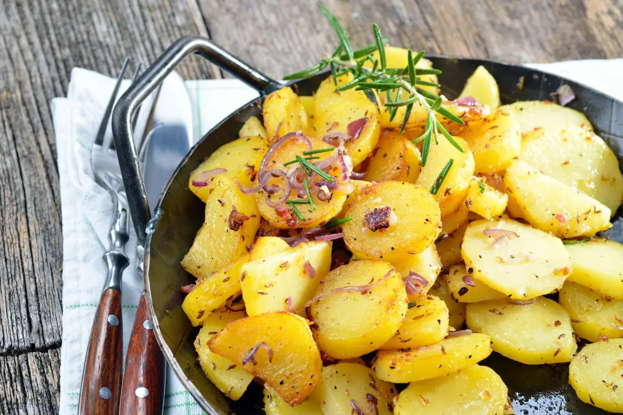 Goldbraune Bratkartoffeln mit Zwiebeln und Rosmarin in einer Pfanne, perfekt, um sie warm zu halten. Dazu liegen Gabel und Messer bereit.