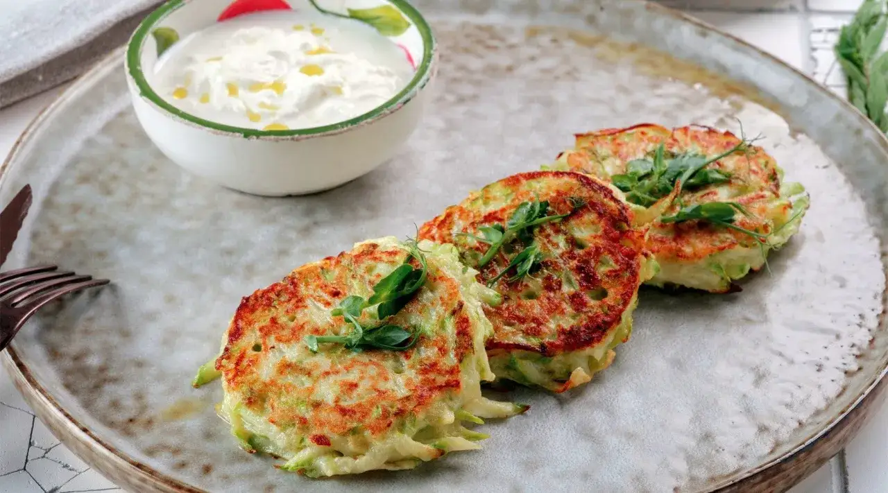 Drei goldbraune Kartoffel-Zucchini-Puffer mit frischen Sprossen garniert, daneben eine Schüssel cremiger Dip.
