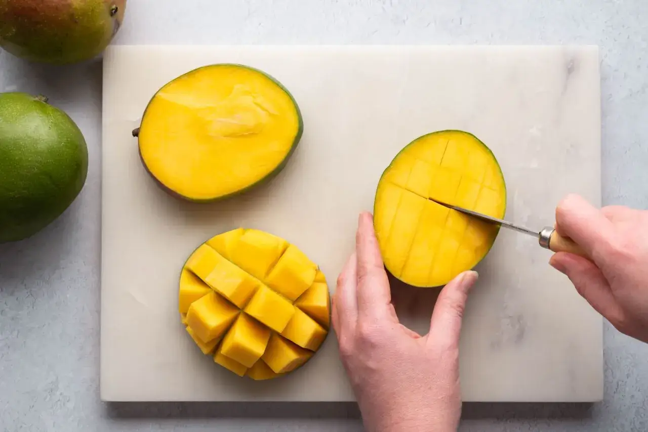 Eine Hand schneidet eine unreife Mango auf einem Marmorbrett in W&uuml;rfel. Daneben liegen zwei weitere Mangoh&auml;lften.