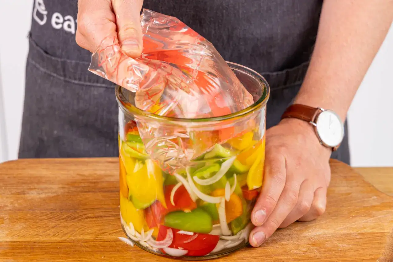 Bunte Paprikastücke und Zwiebeln werden in einem Glas für das Paprika fermentieren vorbereitet. Ein Plastikbeutel wird über das Gemüse gelegt.