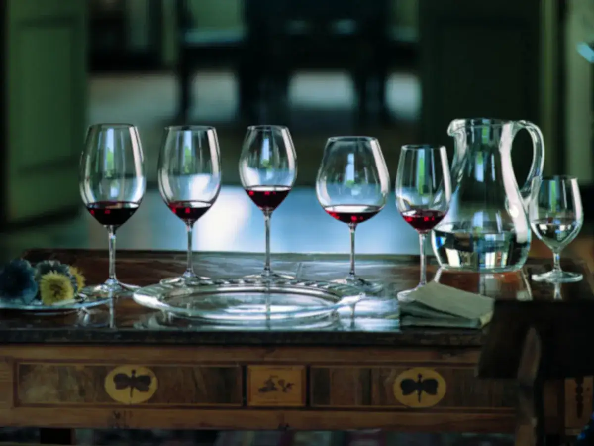 Sechs Gläser mit Rotwein, bereit für eine Verkostung von Cabernet Sauvignon vs. Merlot. Ein Krug mit Wasser und ein paar Dekorationen stehen daneben.