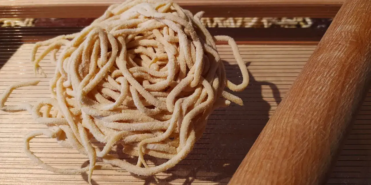 Frische Pasta, alla chitarra, liegt auf einem Holzbrett. Daneben ein Nudelholz.