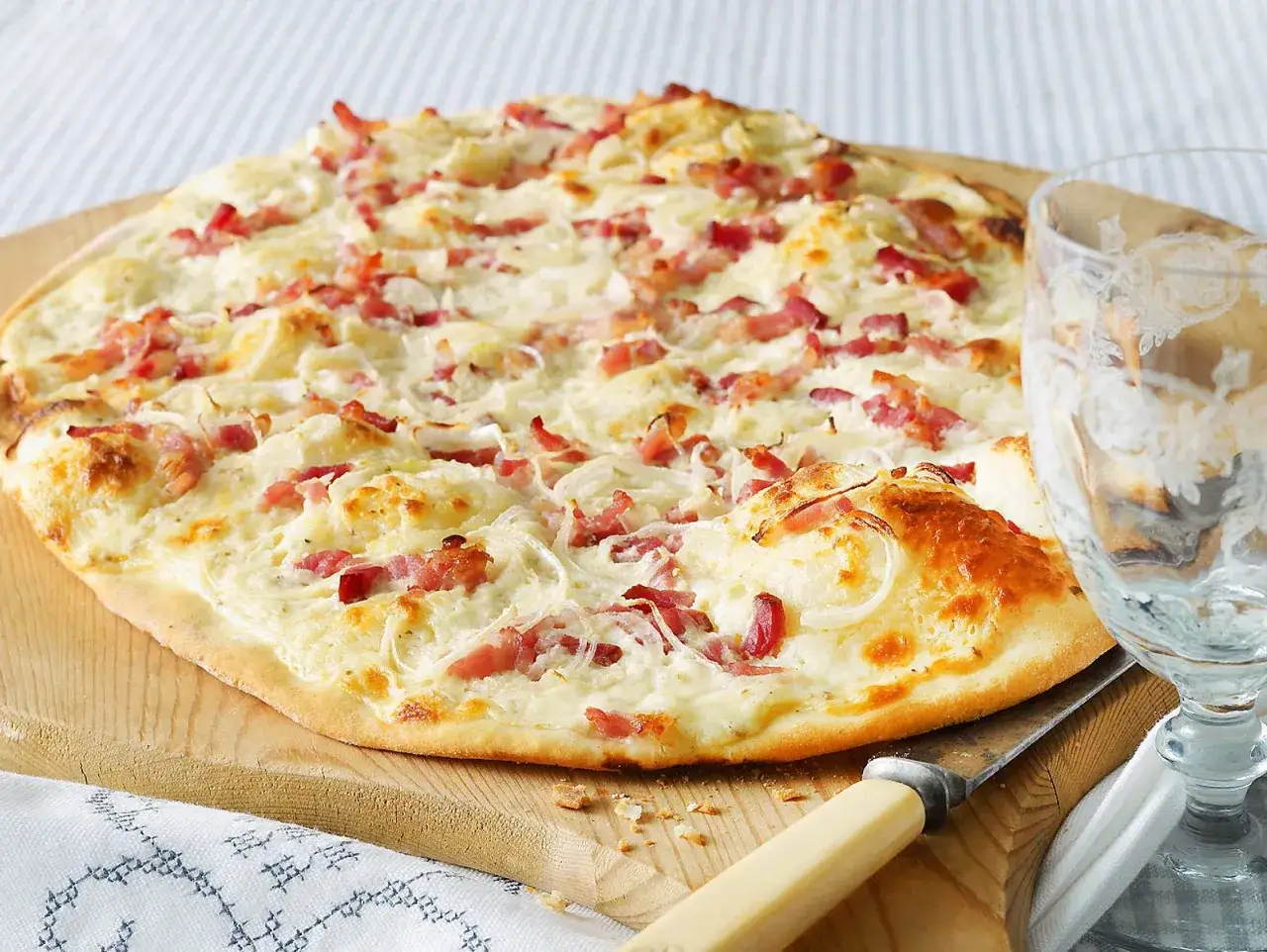 Flammkuchen machen: Ein knuspriger Flammkuchen mit Speck und Zwiebeln auf einem Holzbrett, daneben ein Glas.