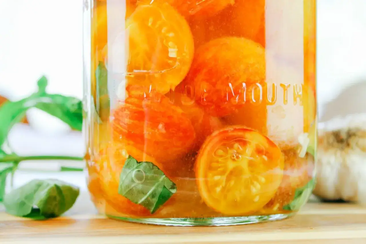 Glas mit leuchtend orangen Tomaten, die fermentieren. Basilikumbl&auml;tter und Knoblauch im Hintergrund.