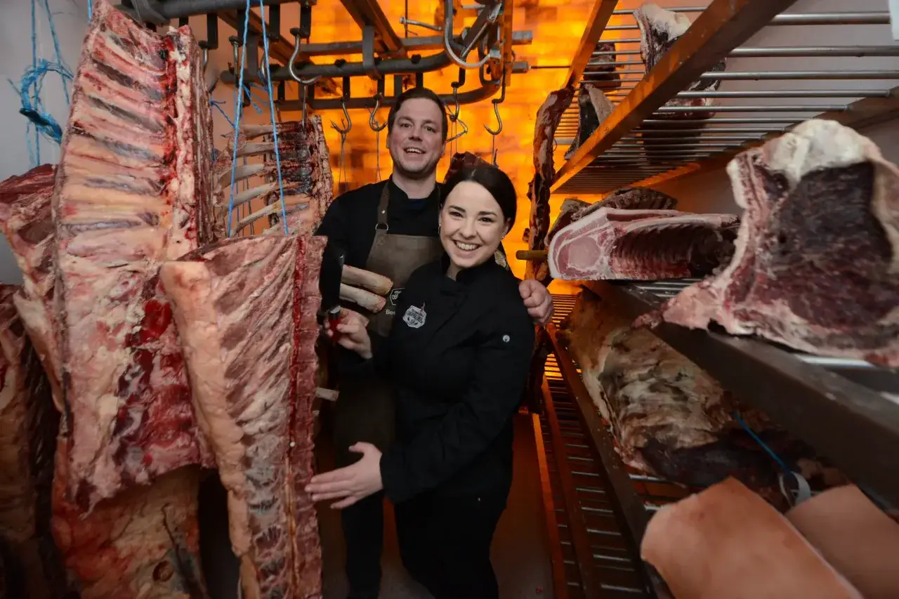 Ein Mann und eine Frau l&auml;cheln inmitten von reifendem Fleisch. Gro&szlig;e Fleischst&uuml;cke h&auml;ngen an Haken und liegen auf Regalen.