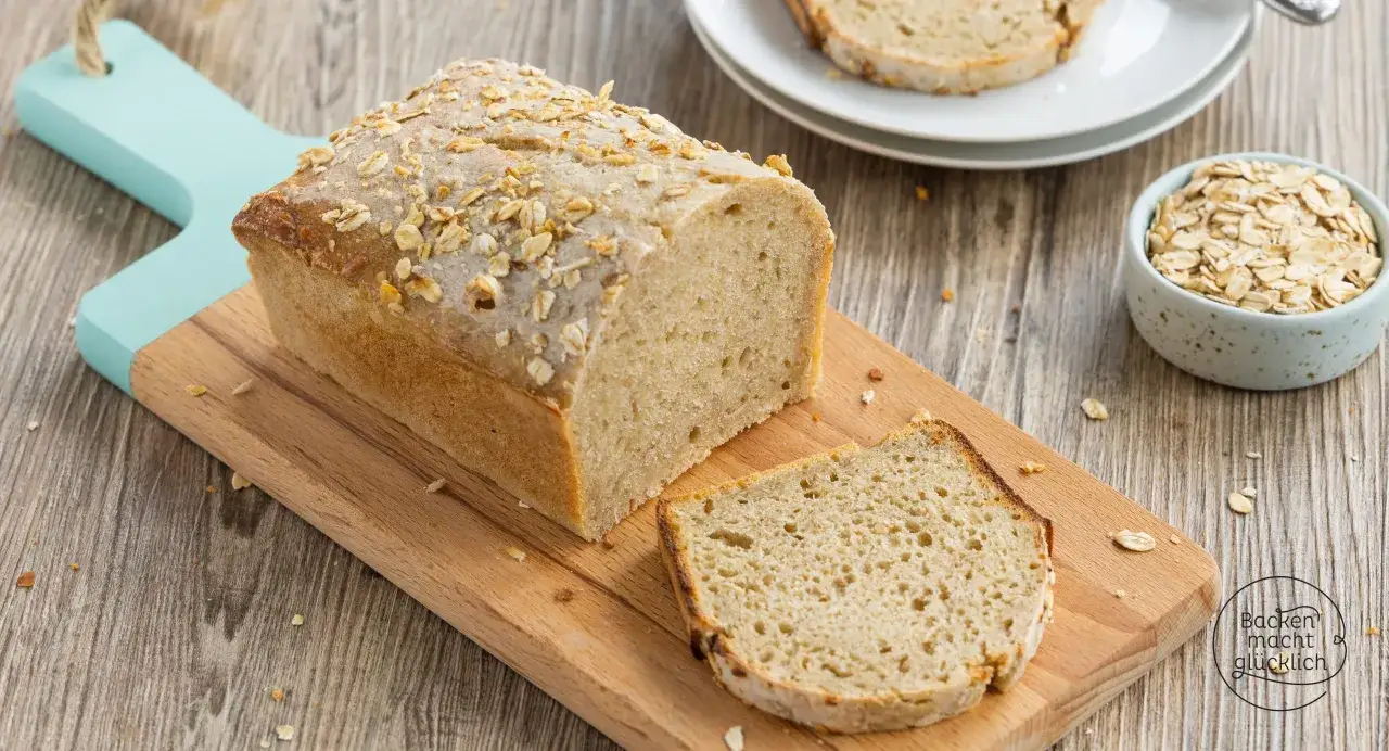 Frisch gebackenes Haferbrot mit Sauerteig, bestreut mit Haferflocken, liegt auf einem Holzbrett. Eine Scheibe ist abgeschnitten.