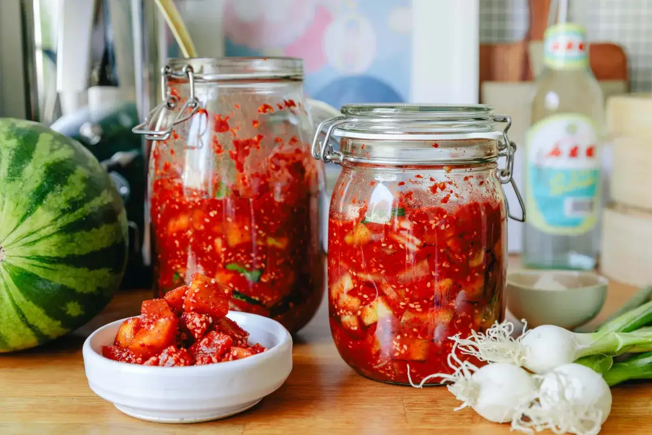 Zwei Gl&auml;ser mit fermentierter Melone, daneben eine Wassermelone, Fr&uuml;hlingszwiebeln und eine Flasche.
